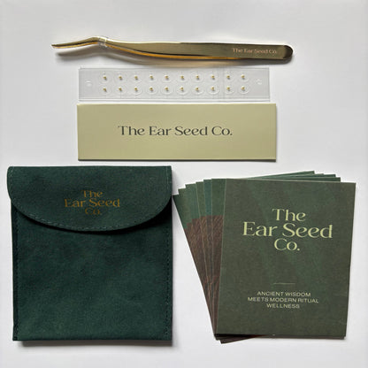The Ear Seed Co. wellness kit laid out on a light background, including a gold ear pick, a clear tray, a beige card, a dark green pouch, and a stack of printed cards or booklet pages.