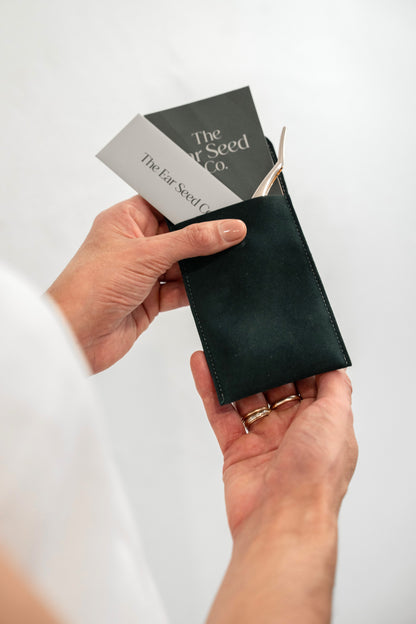 A close-up of a person's hands holding a dark green leather seed packet holder, with seed packets tucked into the pocket. The brand name 'The Far Seed Co.